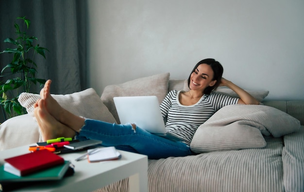 Jonge mooie lachende vrouw met laptop terwijl ze thuis op de grote bank lag en ontspande