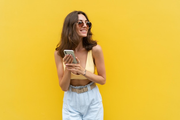 Jonge mooie lachende vrouw met mobiele telefoon Zorgeloos vrouw poseren in de straat in de buurt van gele muur Positief model buitenshuis in zonnebril Blij en opgewonden