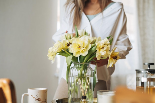 Jonge mooie lachende vrouw veertig jaar met blond lang haar in wit overhemd met boeket gele bloemen in handen in de buurt van raam in licht interieur thuis