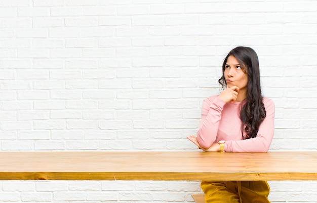 Jonge mooie Latijns-vrouw denken, twijfelachtig en verward voelen, met verschillende opties, zich afvragend welke beslissing om voor een tafel te zitten