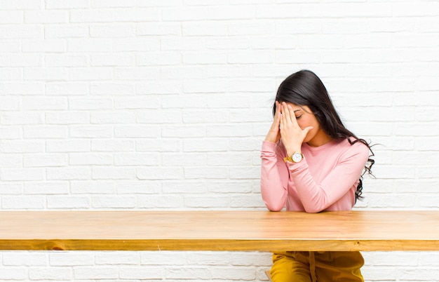 Jonge mooie Latijns-vrouw die de ogen bedekt met handen met een droevige, gefrustreerde blik van wanhoop, huilend, zijaanzicht dat voor een tafel zit