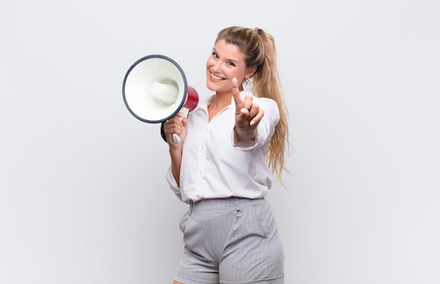 Jonge mooie Latijns-vrouw glimlachend trots en vol vertrouwen waardoor nummer één triomfantelijk poseert, zich een leider voelend met een megafoon tegen de witte muur