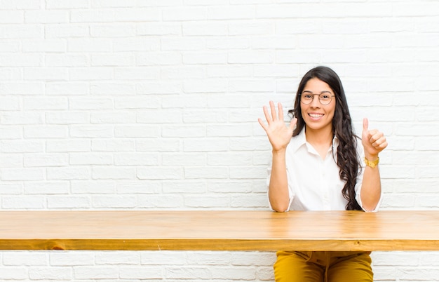 Jonge mooie Latijns-vrouw glimlacht en kijkt vriendelijk, toont nummer zes of zesde met vooruit hand, aftellen zitten voor een tafel