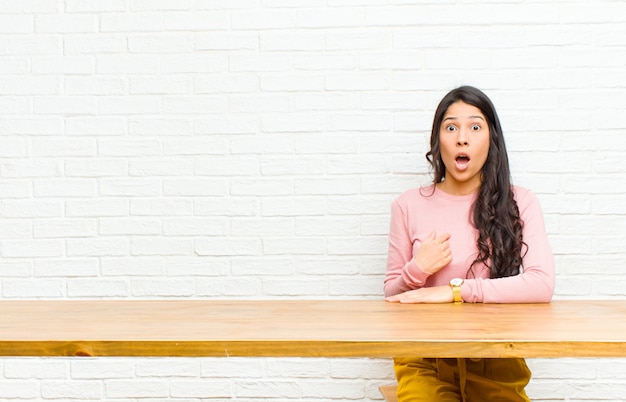 Jonge mooie Latijns-vrouw op zoek geschokt en verrast met wijd open mond, wijzend naar zelf zitten voor een tafel