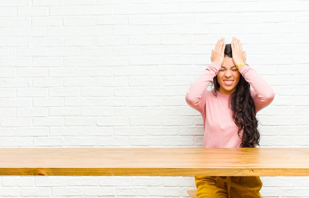 Jonge mooie Latijns-vrouw voelt zich gestrest en angstig, depressief en gefrustreerd met een hoofdpijn, beide handen opheffende zitten voor een tafel