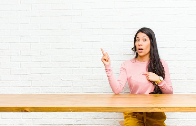 Jonge mooie Latijns-vrouw voelt zich trots en verrast, wijzend op zelfverzekerd, gevoel als succesvolle nummer één zit voor een tafel