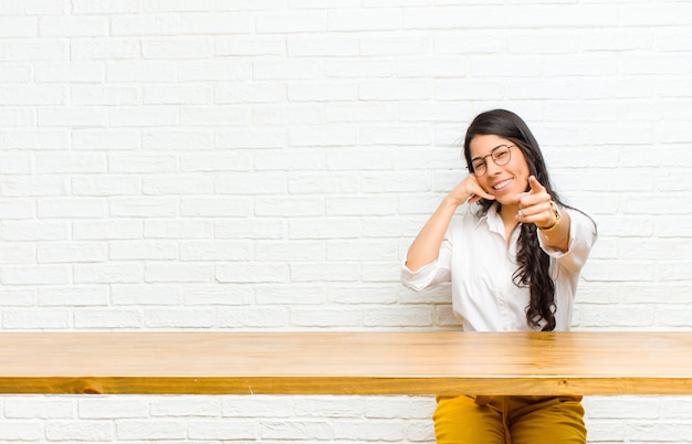 Jonge mooie Latijns-vrouw vrolijk glimlachen en wijzend naar de camera tijdens het bellen u later gebaar, praten over telefoon zit achter een tafel