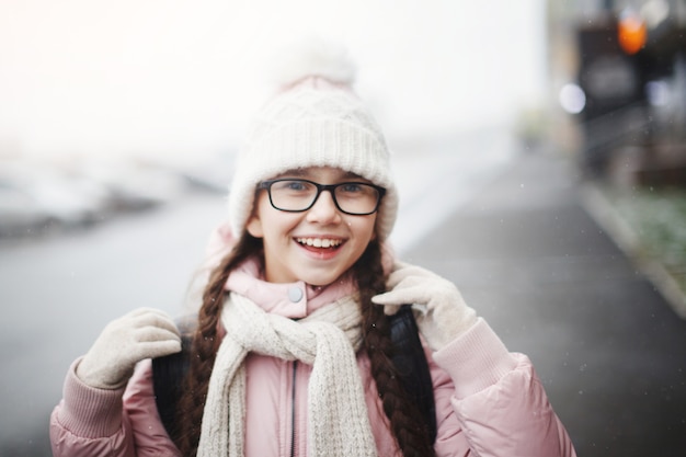 Jonge mooie meisjestiener met lang haar in een bril en in een modieuze witte gebreide muts lacht gelukkig en geniet van het leven buiten in de winter. Detailopname.