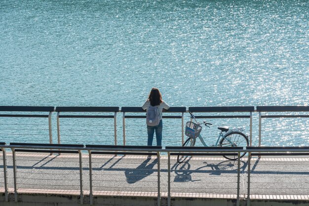 Jonge mooie meisjestribune dichtbij een fiets aan het bekijken het uitzicht op fietspad bij het meer in de ochtend. Actieve mensen. Buitenshuis