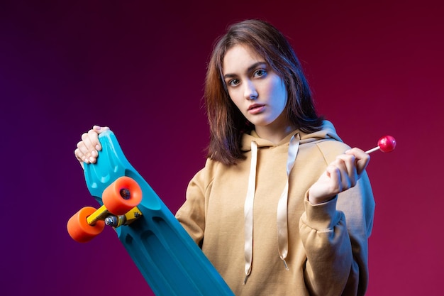 Jonge mooie modieuze hipster meisje gekleed in een hoodie houdt een skateboard in haar handen op een paarse studio achtergrond Eet een lolly