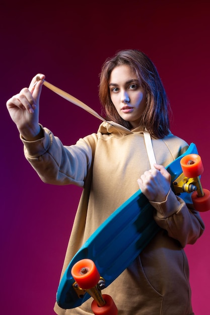 Jonge mooie modieuze hipster meisje gekleed in een hoodie houdt een skateboard in haar handen op een paarse studio achtergrond
