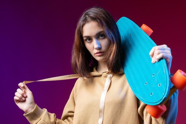 Jonge mooie modieuze hipster meisje gekleed in een hoodie houdt een skateboard in haar handen op een paarse studio achtergrond