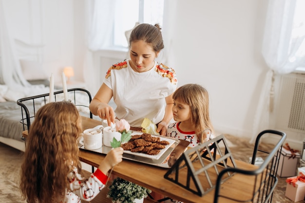 Jonge mooie moeder en haar twee charmante dochtertjes zitten aan tafel en gaan cacao drinken met Marshmallows en koekjes in de gezellige kamer met de nieuwjaarsboom.