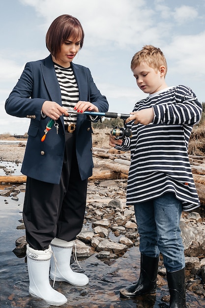 Jonge, mooie moeder en zoon hebben plezier bij het vissen op het meer. Moeder en zoon in gestreepte vesten aan de rivieroever met een hengel.