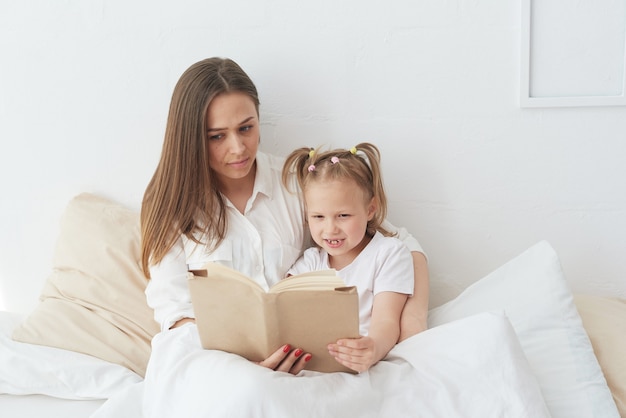 Jonge mooie moeder leest een boek voor aan haar dochter. Gelukkige familie, moeder en dochter lezen een boek terwijl ze in bed liggen, glimlachende oppasmoeder die een grappig verhaal vertelt aan een schattig kleutermeisje.