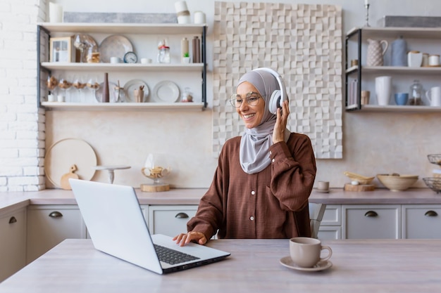 Jonge, mooie moslimstudentenvrouw in hijab die thuis in de keuken zit en een koptelefoon draagt en