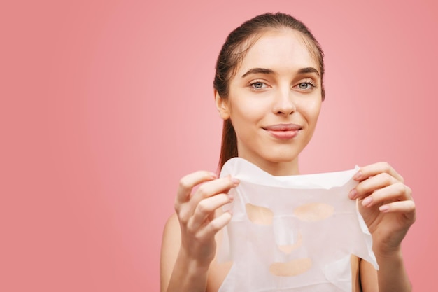 Jonge, mooie naakte vrouw die haar handen afdoet van het gezicht van een wit stoffen cosmetisch masker tegen een roze muurachtergrond