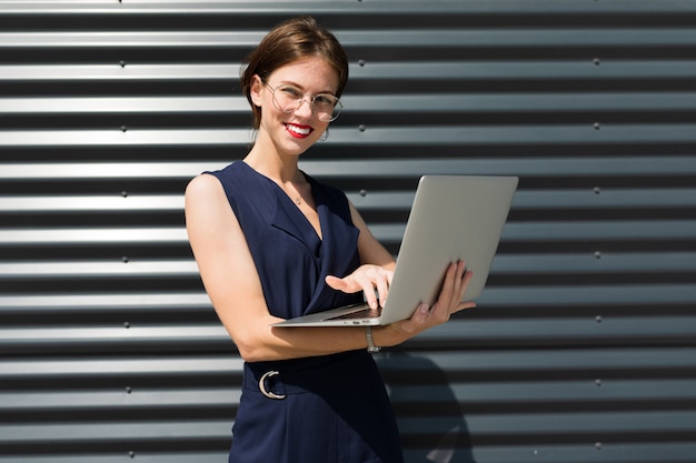 Jonge mooie office vrouw met lichte make-up, rode lippen, bril staat in de buurt van het kantoor en werkt met laptop