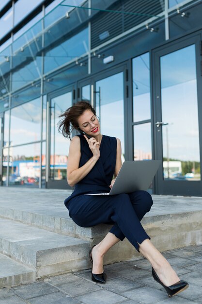 Jonge mooie office vrouw met lichte make-up, rode lippen, bril zit, communiceert met iemand of werkt met laptop