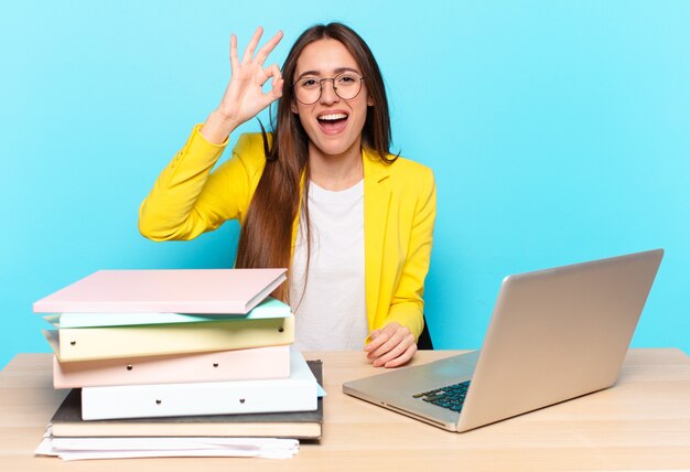 Jonge mooie onderneemster die succesvol en tevreden voelt, glimlachend met wijd open mond, goed teken met hand maakt