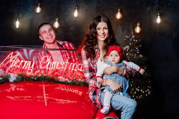 Jonge mooie ouders houden hun kleine schattige dochter in hun armen met plezier in de buurt van retro auto in de studio. kerst familie look. nieuwjaars landschap.