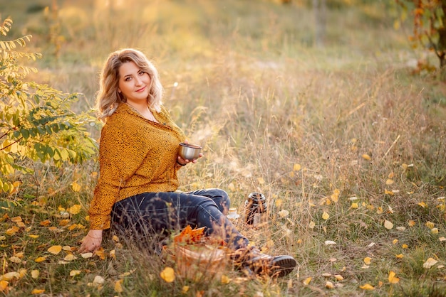 Jonge mooie plussize vrouw in een oranje shirt en spijkerbroek zit in een herfstpark thee drinken en ontspannen op een picknick