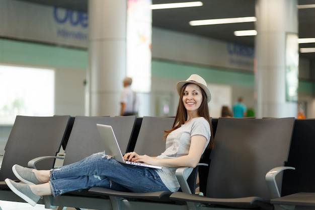 Jonge mooie reizigerstoeristenvrouw met hoed die aan laptop werkt terwijl ze wacht in de lobby op de internationale luchthaven