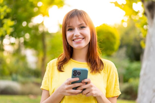 Jonge mooie roodharige vrouw buitenshuis die naar de camera kijkt en glimlacht tijdens het gebruik van de mobiel