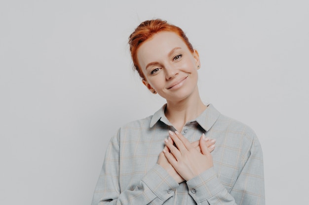 Jonge mooie roodharige vrouw die gevouwen handen op de borst houdt en zich dankbaar en dankbaar voelt