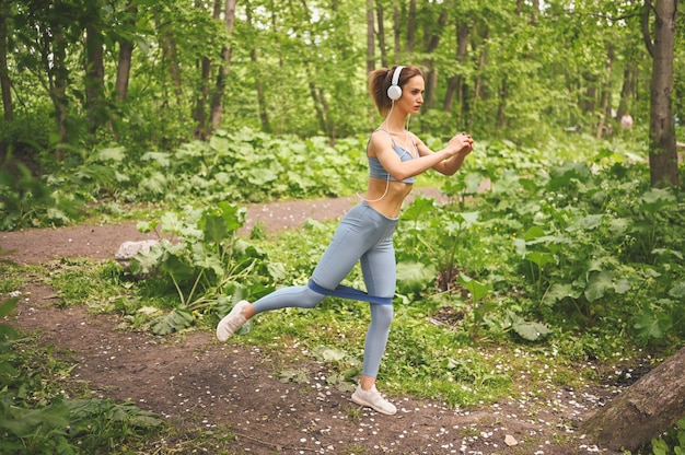 Jonge mooie slim fit meisje in blauwe sportkleding met grote witte koptelefoon doen oefening met fitness training gom in het park