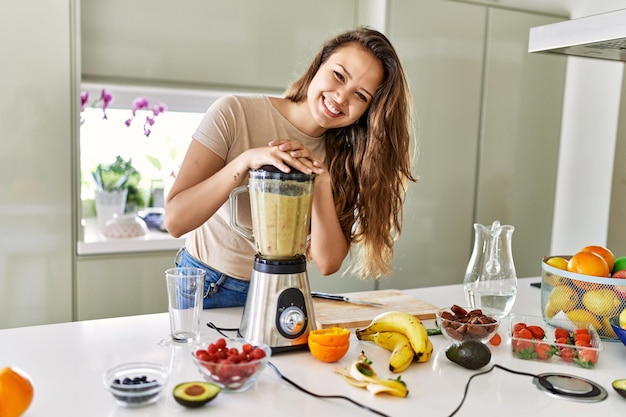 Jonge mooie spaanse vrouw die groentesmoothie voorbereidt met blender in de keuken