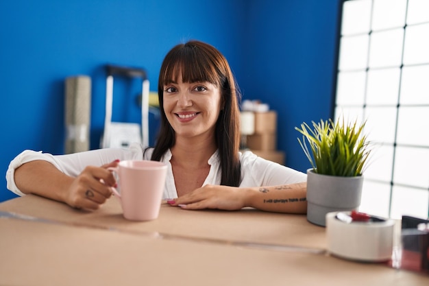 Jonge mooie Spaanse vrouw die koffie drinkt leunend op pakket in nieuw huis