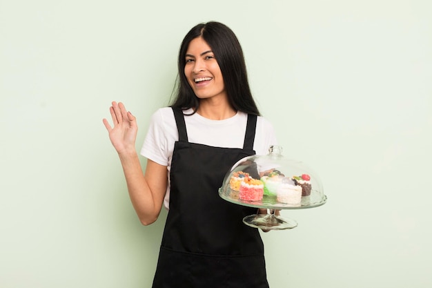 Jonge, mooie Spaanse vrouw die lacht en vrolijk zwaait met de hand, verwelkomt en begroet je chef-kok met taartenconcept