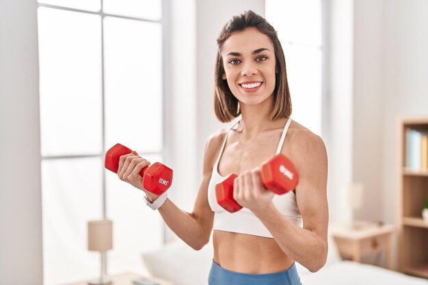 Jonge mooie spaanse vrouw die lacht zelfverzekerd met behulp van halters die trainen in de slaapkamer