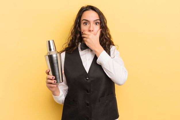 Jonge mooie Spaanse vrouw die mond bedekt met handen met een geschokt barman en cocktailconcept