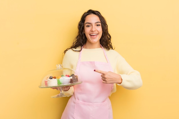 Jonge mooie Spaanse vrouw die opgewonden en verrast kijkt en wijst naar het concept van de chef-kok en gebak
