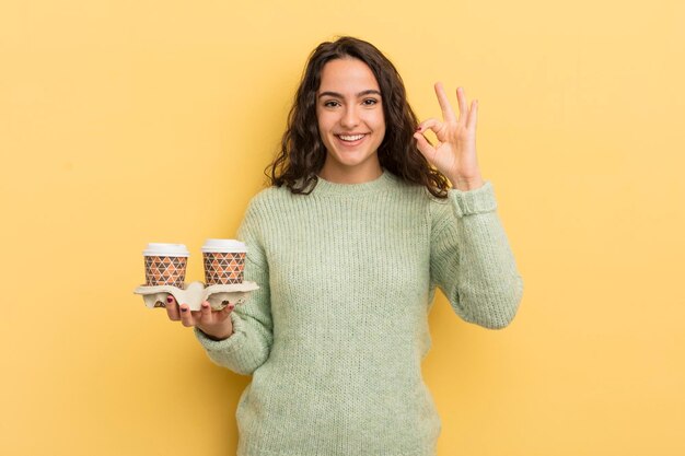 Jonge mooie Spaanse vrouw die zich gelukkig voelt en goedkeuring toont met een goed gebaar om koffieconcept weg te nemen