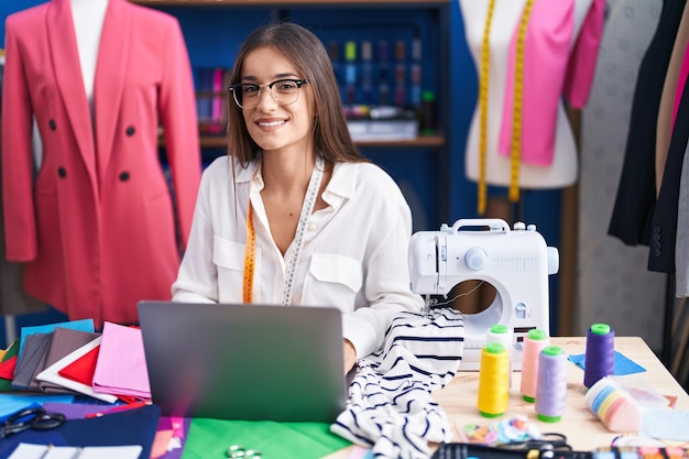 Jonge mooie spaanse vrouw kleermaker glimlachend zelfverzekerd met behulp van laptop in kledingfabriek