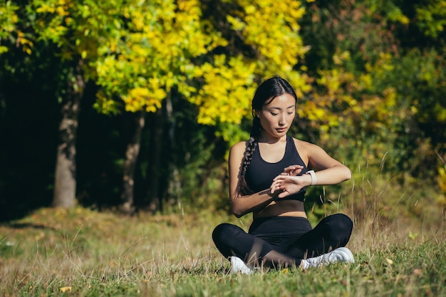 Jonge mooie sport meisje aziatische vrouw mediteren in park, zitten lotus pose beoefenen yoga mat buitenshuis kijken slim horloge op moderne track met behulp van gebruik smartwatch fitness armband resultaat controleren