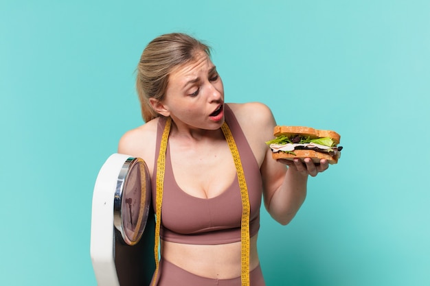 Jonge mooie sportvrouw die twijfelt of een onzekere uitdrukking heeft en een weegschaal en een broodje vasthoudt