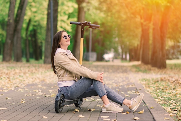 Jonge, mooie, stijlvolle vrouw die op een elektrische scooter rijdt in het ecologisch transport van het herfstpark