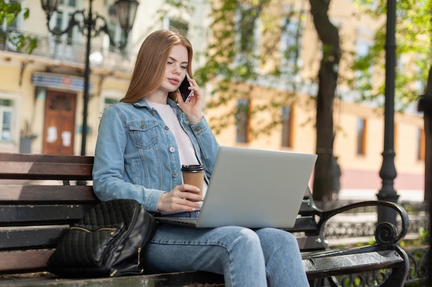 Jonge mooie studente of freelancer die laptop gebruikt en aan de telefoon praat terwijl ze op een bankje in de stad zit