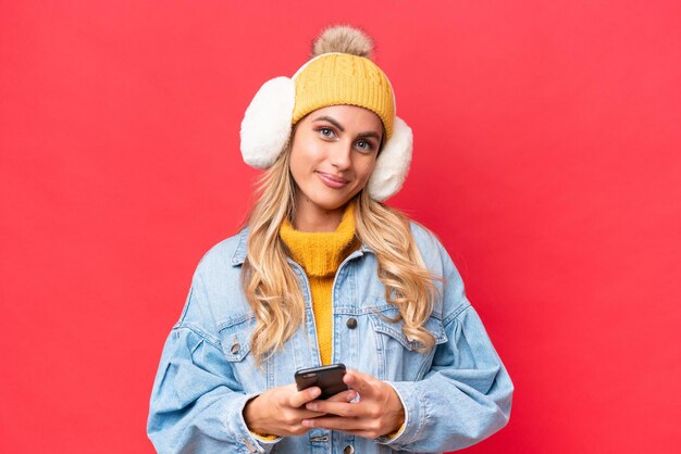 Jonge mooie Uruguayaanse vrouw met wintermoffen geïsoleerd op rode achtergrond achtergrond die een bericht verzendt met de mobiel