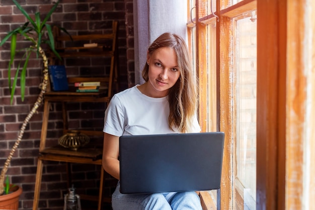 Jonge mooie vrouw bij raam zitten en werken met laptop.