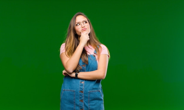 Jonge mooie vrouw denken, twijfelachtig en verward voelen, met verschillende opties, zich afvragend welke beslissing te nemen tegen groene muur