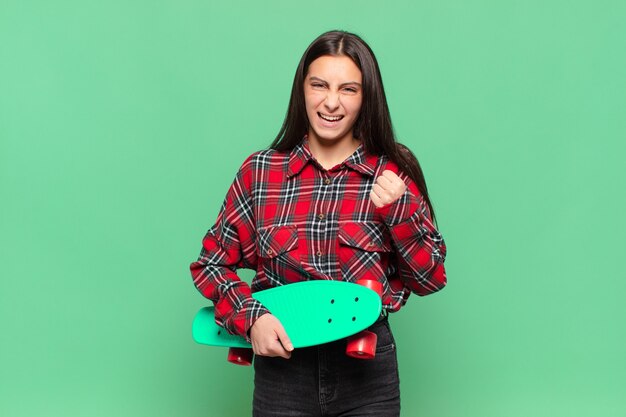 Jonge mooie vrouw die agressief schreeuwt met een boze uitdrukking of met gebalde vuisten om succes te vieren. skate board concept