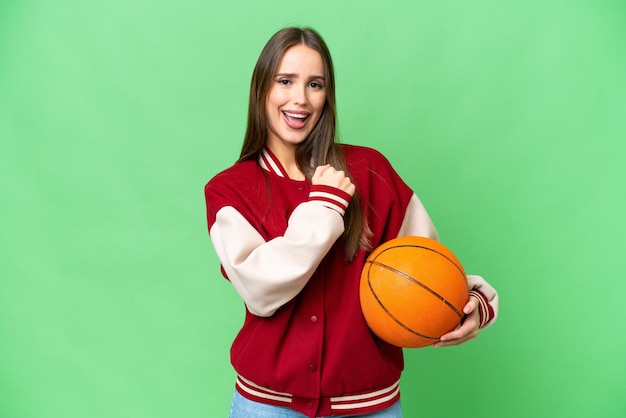 Jonge mooie vrouw die basketbal speelt over geïsoleerde chroma key achtergrond die een overwinning viert