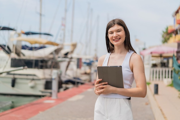 Jonge mooie vrouw die buitenshuis een tablet vasthoudt met een gelukkige uitdrukking