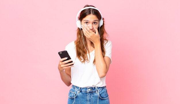 Jonge mooie vrouw die de mond bedekt met handen met een schok met een koptelefoon en een smartphone