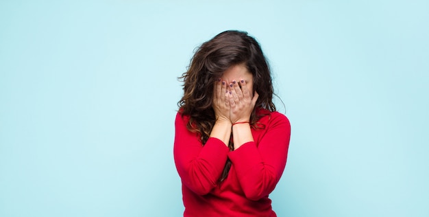 Jonge mooie vrouw die droevig, gefrustreerd, zenuwachtig en depressief voelt, die gezicht behandelt met beide handen, die tegen blauwe muur schreeuwt